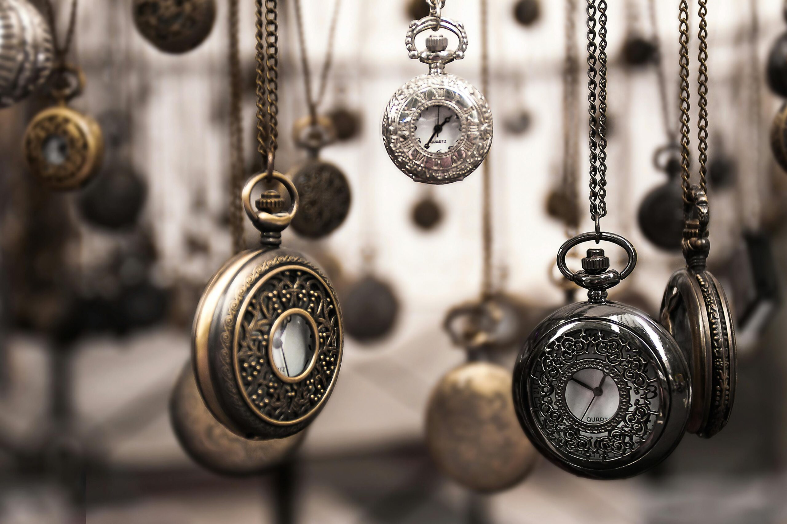 Close-up of antique pocket watches with intricate designs hanging in a blurred vintage setting.